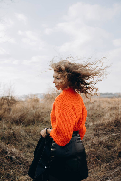 Against the background of autumn park smiles beautiful smile curly brunette. young curly girl in an orange sweater against the backdrop of autumn nature - Zdjęcie, obraz