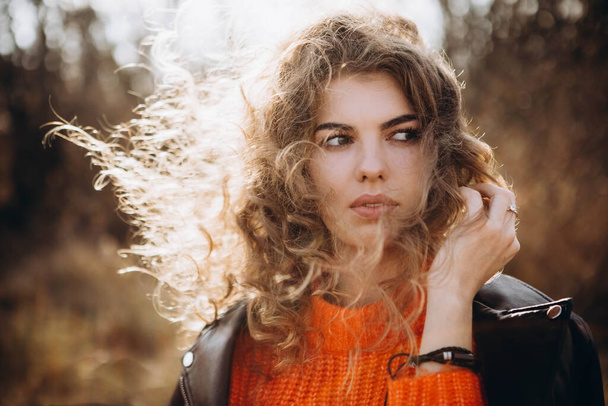 На фоне осеннего парка улыбается красивая кудрявая брюнетка. молодая кудрявая девушка в оранжевом свитере на фоне осенней природы - Фото, изображение