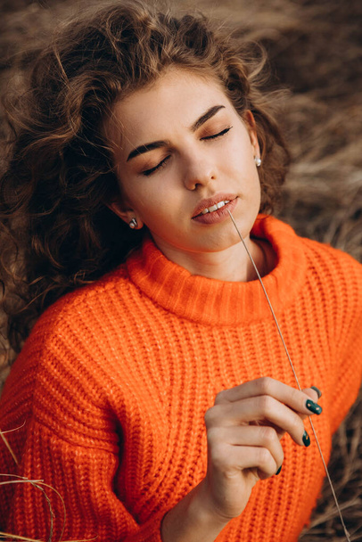 Against the background of autumn park smiles beautiful smile curly brunette. young curly girl in an orange sweater against the backdrop of autumn nature - Photo, Image