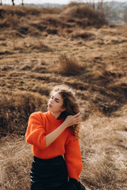 En el fondo del parque de otoño sonríe hermosa sonrisa morena rizada. joven chica rizada en un suéter naranja contra el telón de fondo de la naturaleza de otoño - Foto, Imagen