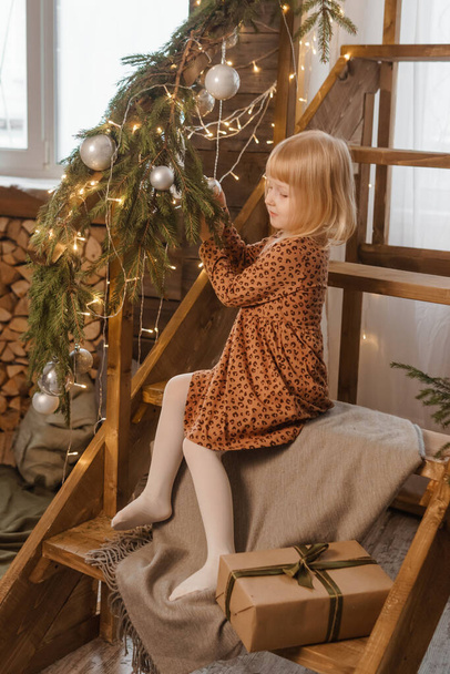 Ein kleines blondes Mädchen sitzt auf einer Holztreppe in einem skandinavischen Interieur. - Foto, Bild