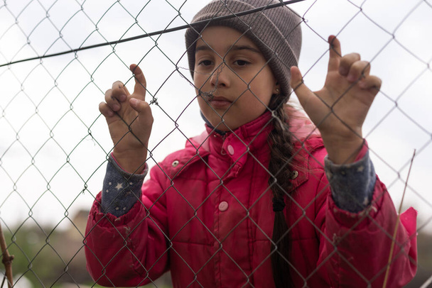 Ένα μικρό κορίτσι με ένα θλιμμένο βλέμμα πίσω από ένα μεταλλικό φράχτη. Κοινωνικό πρόβλημα προσφύγων και αναγκαστικών μεταναστών. - Φωτογραφία, εικόνα
