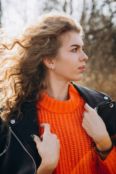 Contra o fundo do parque de outono sorri belo sorriso encaracolado morena. jovem garota encaracolado em uma camisola laranja contra o pano de fundo da natureza outono - Foto, Imagem