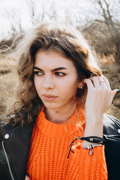 На фоне осеннего парка улыбается красивая кудрявая брюнетка. молодая кудрявая девушка в оранжевом свитере на фоне осенней природы - Фото, изображение