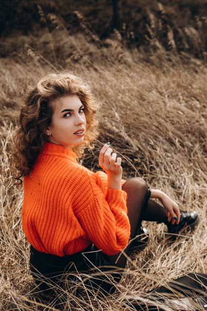 Against the background of autumn park smiles beautiful smile curly brunette. young curly girl in an orange sweater against the backdrop of autumn nature - Foto, immagini
