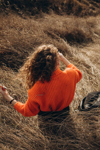 Against the background of autumn park smiles beautiful smile curly brunette. young curly girl in an orange sweater against the backdrop of autumn nature - Foto, immagini