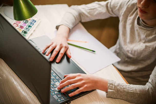 Το αγόρι γράφει την εργασία σε ένα κενό φύλλο που βρίσκεται κοντά στο laptop. Teenager κάνει την εργασία για online μαθήματα με σχέδιο. Αποεστίαση. - Φωτογραφία, εικόνα