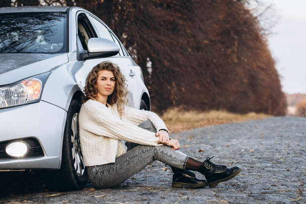 joven chica rizada en un suéter blanco contra el telón de fondo de la naturaleza otoñal y su propio coche. Foto de alta calidad - Foto, imagen