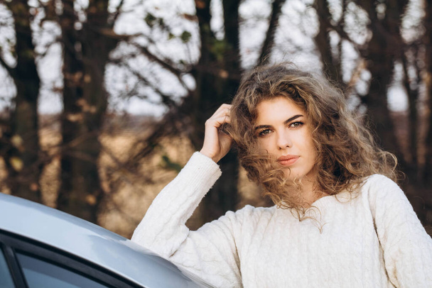 joven chica rizada en un suéter blanco contra el telón de fondo de la naturaleza otoñal y su propio coche. Foto de alta calidad - Foto, imagen