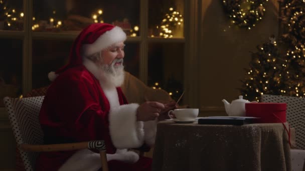 Happy Santa Claus sedí u stolu na dvorku a čte dopis, usmívá se a tančí, pije čaj, rád zjistí, že děti si přeje zblízka zpomalit, zpomalit. - Záběry, video
