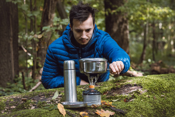 Man in a blue down jacket boils water for tea and food on a portable stove and in a stainless steel ultralight pot in the middle of the wilderness. Hiking lifestyle. - Fotoğraf, Görsel
