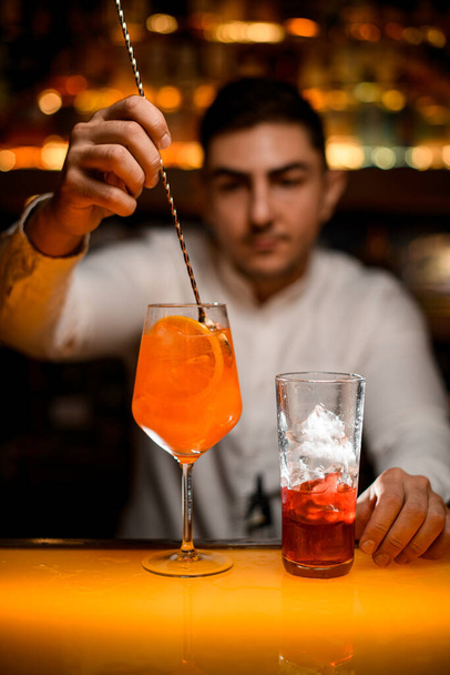 hand of male bartender gently stirs cocktail with orange slices in glass on bar counter with spoon. Close-up. - Φωτογραφία, εικόνα