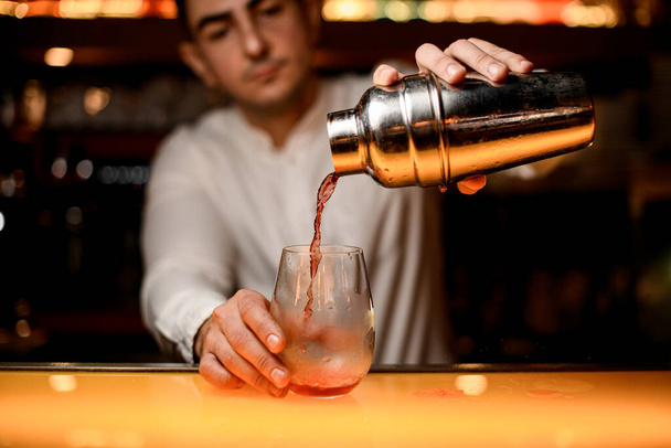 hand of male bartender carefully holds steel shaker and pours cocktail into transparent glass on the bar counter - Fotó, kép