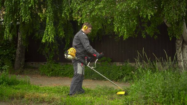 "Молодой человек из спецслужб стрижет газон бензиновым триммером. Человек в очках и наушниках с газонокосилкой в руках." - Фото, изображение
