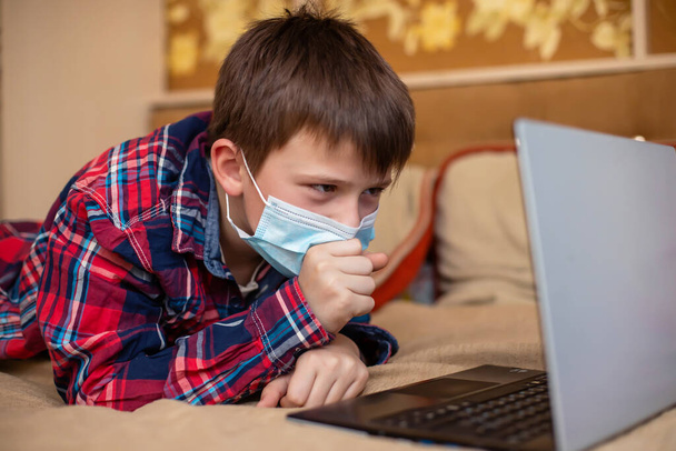 保護医療マスクの10代の少年が最初に咳をする。子供はノートパソコンの近くのベッドで遠隔授業をしています。コロナウイルス隔離中のオンライントレーニング - 写真・画像