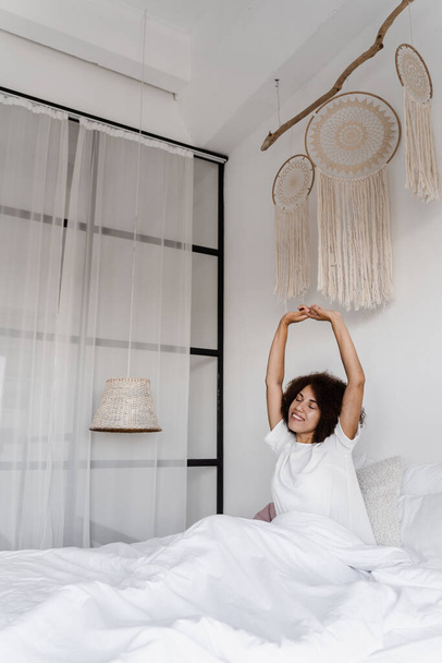 Mujer afroamericana despierta y disfruta de buenos días. Chica africana en pijama esteretizando en la cama y levantando las manos en casa - Foto, Imagen