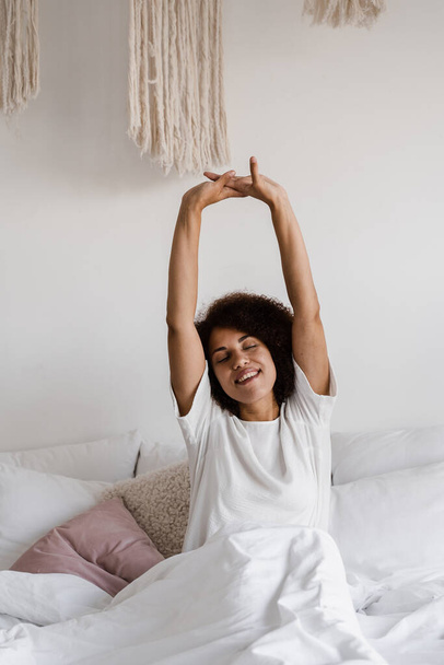 Afroamerikanerin wacht auf und genießt guten Morgen. Afrikanisches Mädchen im Schlafanzug liegt im gemütlichen Bett und hebt zu Hause die Hände - Foto, Bild