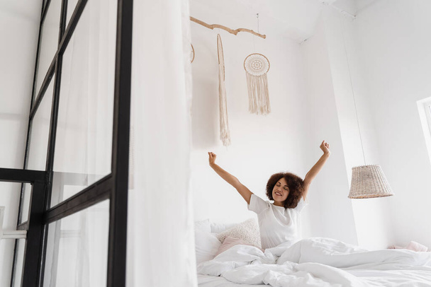 アフリカ系アメリカ人の女性が目を覚ますとおはようございます。アフリカの女の子でパジャマstertchingで居心地の良いベッドと上げて手アップで家 - 写真・画像