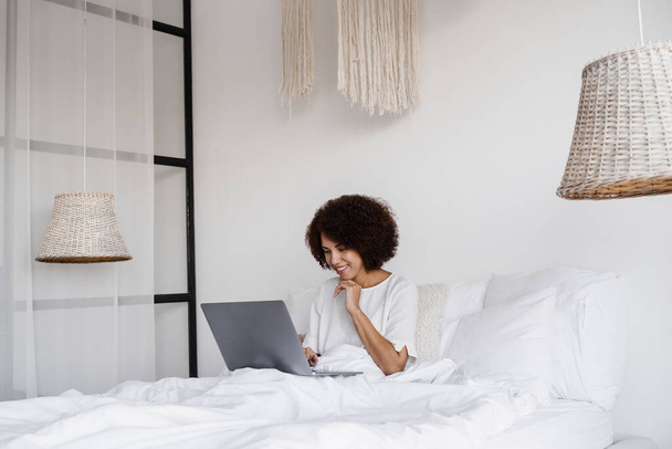 アフリカ系アメリカ人の女子学生は、自宅で居心地の良い朝にベッドの上でノートパソコンで勉強します。家からの距離学習 - 写真・画像