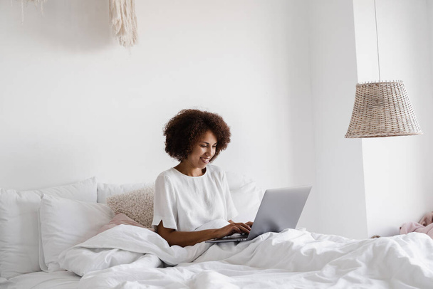 African girl manager with laptop have online video meeting with colleagues and business partners in cozy bed at home in the morning. Team discussion trends and tasks - Фото, изображение