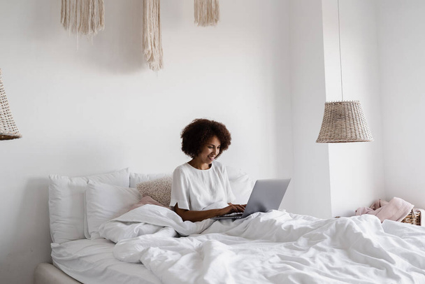 Chica africana gerente con el ordenador portátil tienen video en línea reunión con colegas y socios de negocios en la acogedora cama en casa por la mañana. Tendencias y tareas del equipo de discusión - Foto, imagen