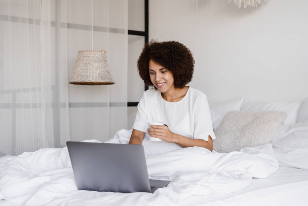 Happy african girl with laptop drinking cup of coffee, watching online movie and surfing internet in bedroom at home. Cozy morning with laptop of african american woman - Foto, imagen