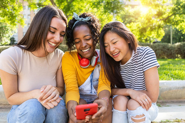 Three young multiracial women friends using mobile phone sitting outdoors - Female friendship concept with millennial people sharing social media content on smart phone device - Foto, imagen