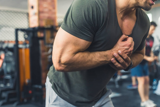 Unrecognizable caucasian male body-builder in greenish gray t-shirt tightening his arm muscles at gym. Blurred background. High quality photo - Foto, immagini
