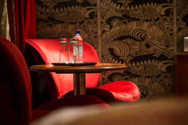 Shot of water with two glasses on table in an empty restaurant with red seats and wall paper. - Foto, Imagen