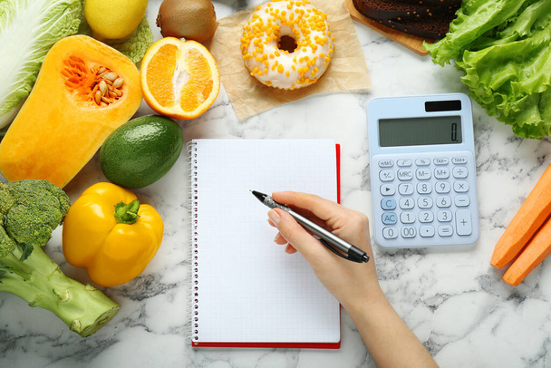 Woman calculating calories at white marble table with food products, top view. Weight loss concept - Foto, imagen