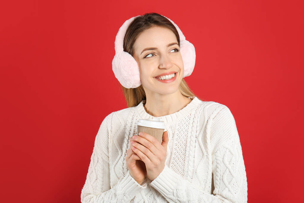 Happy woman with cup of drink wearing warm earmuffs on red background - Fotó, kép