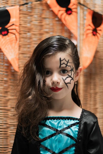 Portrait of a beautiful Caucasian girl in a witch dress with Halloween makeup on her face stands against the background of a wicker fence with holiday flags and looking at the camera, close-up side view. - Foto, Bild