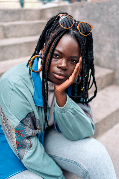 Expressive young african woman looking at camera outdoors and posing. She is sitting in stairs in the city. - Fotó, kép