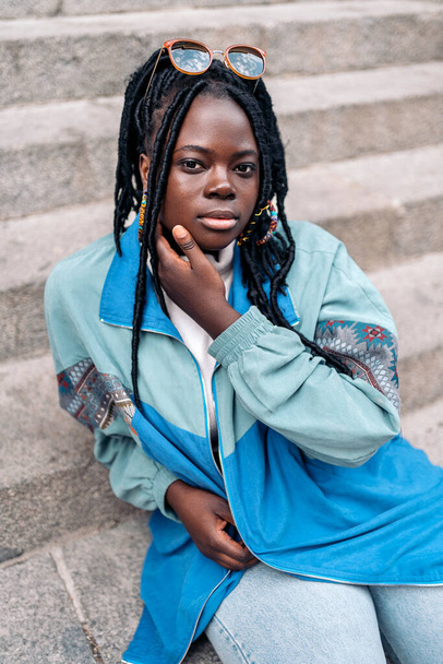 Expressive young african woman looking at camera outdoors and posing. She is sitting in stairs in the city. - Foto, afbeelding