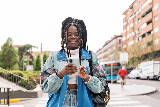 Happy african woman with cool braids walking in the street and typing in her mobile phone. - Φωτογραφία, εικόνα