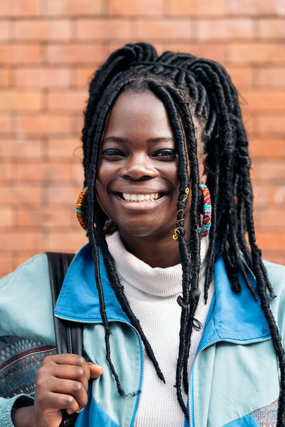 Happy and young african woman with cool braids smiling and looking at camera. She is wearing a backpack. - Valokuva, kuva