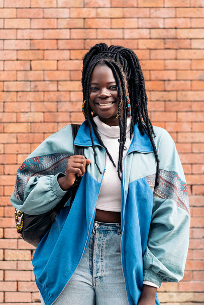 Happy and young african woman with cool braids smiling and looking at camera. She is wearing a backpack. - Foto, afbeelding