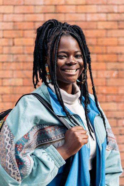 Happy and young african woman with cool braids smiling and looking at camera. She is wearing a backpack. - Foto, Imagem