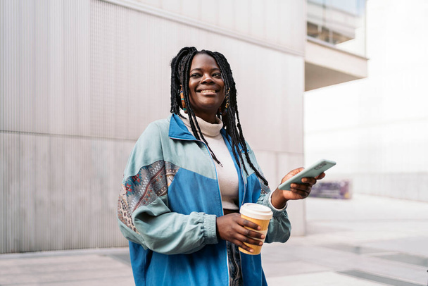 Beautiful african woman walking in the street using her phone and holding coffee cup. - Foto, Bild