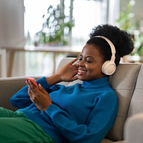 Happy African American kobieta w słuchawkach słuchających muzyki za pomocą telefonu komórkowego, aby wybrać listę odtwarzania w popularnej aplikacji online. Uśmiechnięta młoda dziewczyna wieku leży na kanapie domu i podcast przesłuchanie lub audiobook - Zdjęcie, obraz