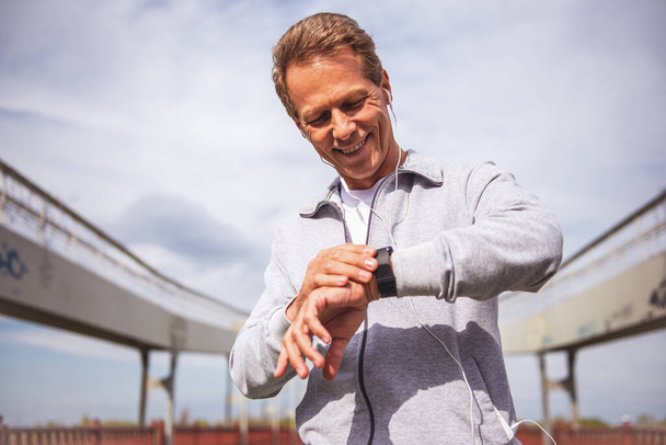 Красивий чоловік середнього віку в спортивній формі і навушники дивиться на його годинник і посміхається під час ранкового бігу
 - Фото, зображення