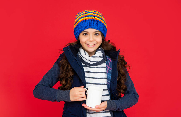cheerful teen girl drink warm tea on red background. portrait of child with coffee cup. drinking cocoa and milk. kid in puffer waistcoat and hat. - Fotó, kép