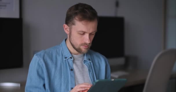 Enthousiaste ingénieur logiciel masculin barbu debout sur le lieu de travail dans le bureau de travail à l'aide d'une tablette. Jeune homme beau en chemise denim, avec des moniteurs d'ordinateur sur fond - Séquence, vidéo