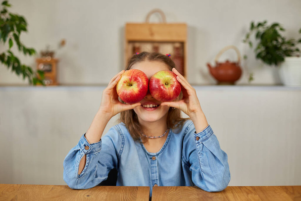 Happy emotional little girl holding red an apple in hands covering eyes at home, smiling children loving fruit healthy snack, vegetarian nutrition for kids, vitamins for children. - Photo, image