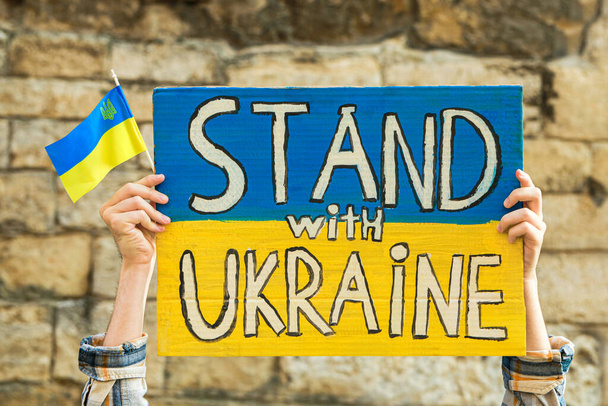 Woman holding poster in colors of national flag and words Stand With Ukraine against brick wall outdoors, closeup - Fotoğraf, Görsel
