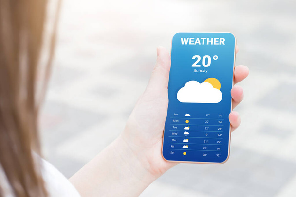 The girl checks the weather forecast in a mobile application. A woman's hand holds a mobile phone and looks at the weather forecast for the day on a park with a trees background - Photo, Image