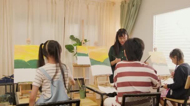 A female Asian teacher teaches and demonstrates to the children on acrylic color picture painting on canvas in art classroom, creatively learning with skill at the elementary school studio education. - Footage, Video