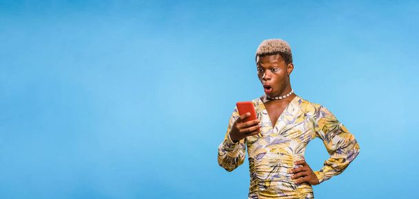 Africaine androgyne personne en robe élégante souriant et envoyer un message texte sur smartphone sur fond bleu - Photo, image