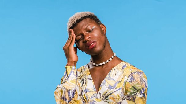 Afrikanische Transgender-Person in stylischem Kleid reibt sich die Stirn und zwinkert, während sie an Migräne vor blauem Hintergrund leidet - Foto, Bild