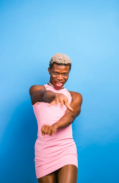 Modèle androgyne africain en robe rose élégante regardant la caméra et dansant sur fond bleu - Photo, image